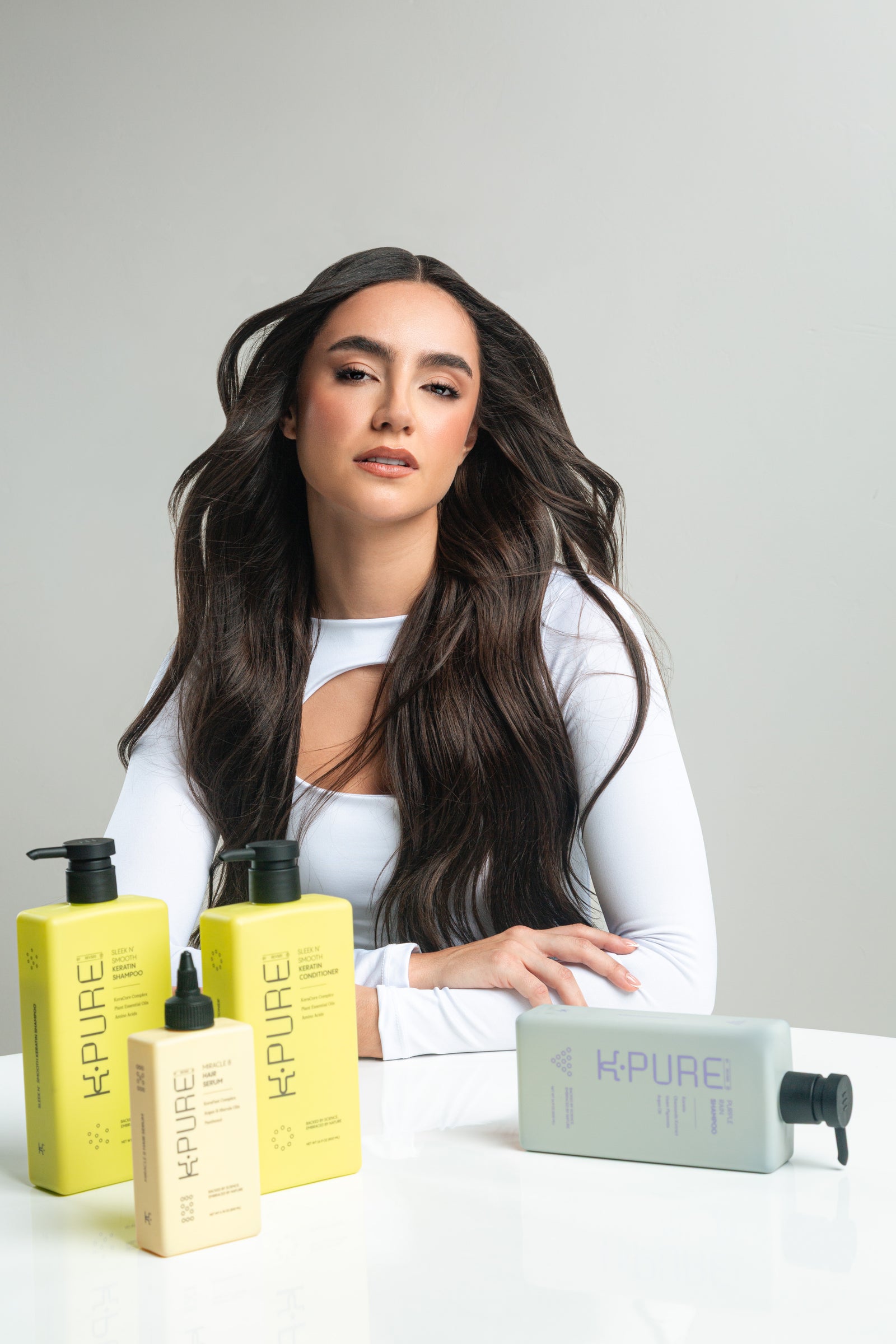 Woman with long hair sitting behind Kpure hair care products on a white background